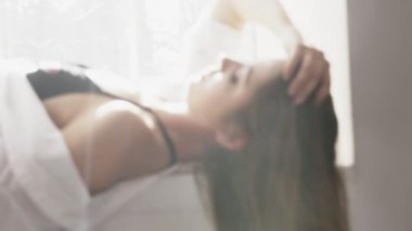 Genç güzel seksi beyaz kızı beyaz gömlek ve siyah iç çamaşırı, yatıyor pencerenin yanında seyir fotoğraf makinesi, portre, günaha, seksi, bulanık arka planda beyaz güneş ışığı. 60 fps