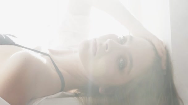 Junge schöne sexy kaukasische Mädchen in weißem Hemd und schwarzer Dessous, liegt am Fenster und blickt in die Kamera, Porträt, Versuchung, sexy, weißes Sonnenlicht im Hintergrund. 60 fps — Stockvideo