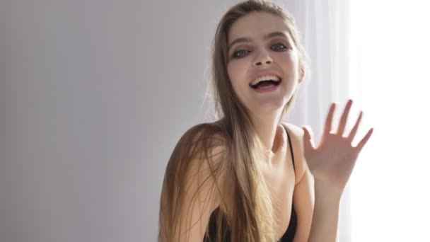 Молодая красивая сексуальная девушка в черном нижнем белье показывает приветственный знак, машет рукой, улыбается, загорает у окна, шторка 50 кадров в секунду — стоковое видео