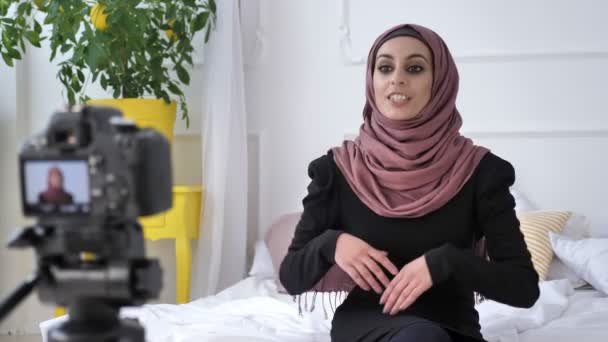 Ung vacker indisk flicka i hijab blogger talar på kameran, leende, gestikulerande, vitt rum, hem komfort i bakgrunden. Stäng upp 50 fps — Stockvideo