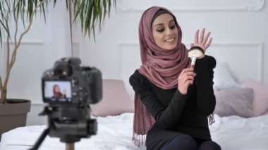 Gülümseyen, kameraya konuşurken, bir yeni satın alma, makyaj fırça, makyaj kavramı, ev konfor arka planda gösterilen genç güzel Hint kızı kameraya konuşurken hijab blogger. 60 fps