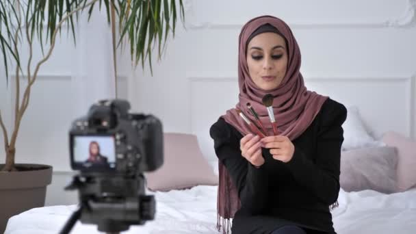 Menina indiana bonita nova no blogueiro hijab falando na câmera, sorrindo, falando para a câmera, mostrando uma nova compra, três pincéis de maquiagem, conceito de maquiagem, conforto em casa no fundo. 60 fps — Vídeo de Stock