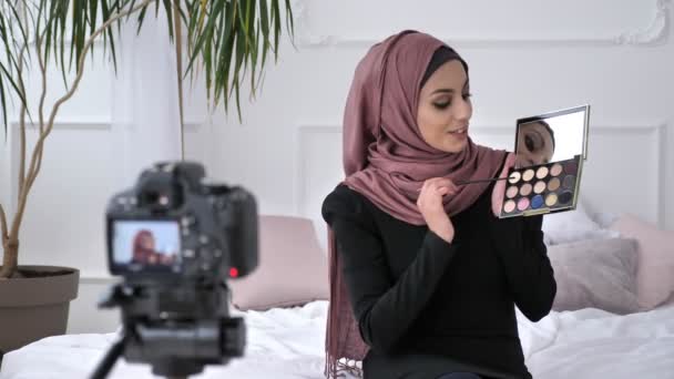 Молодая красивая индийская девушка в хиджабе блоггер говорит на камеру, улыбаясь, показывая новую покупку, косметика, поддон тени для глаз, домашний комфорт на заднем плане. 60 кадров в секунду — стоковое видео