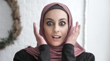 Hijab gülümseyen içinde genç Hintli kızı gülen, şaşırttı, yanaklarını elleriyle, güneş ışığı, pencere beyaz yakınındaki 50 fps arka plan dokunuşlar
