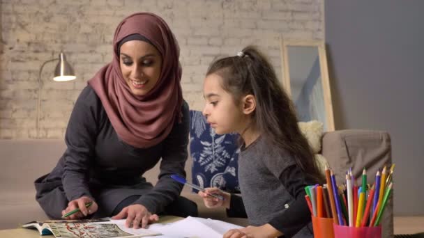 Joven hermosa madre en hijab haciendo la tarea con su hija pequeña, entrenamiento, tarea, comodidad en el hogar en el fondo, lápices de colores 50 fps — Vídeos de Stock