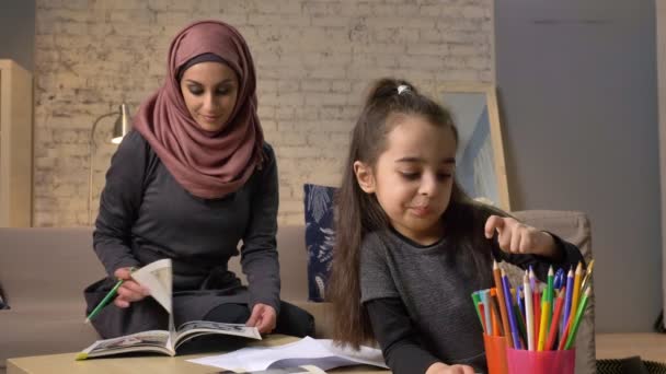 Une jeune belle mère dans un hijab feuilletant un livre, une petite fille choisit des crayons de couleur, devoirs, confort à la maison en arrière-plan, crayons de couleur 50 fps — Video