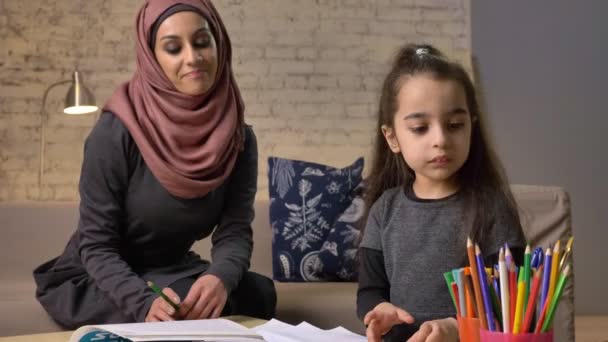 Jeune belle mère en hijab fait des leçons, petite fille choisit des crayons de couleur, dessine, coloriage des enfants, confort à la maison en arrière-plan, crayons de couleur 50 images par seconde — Video