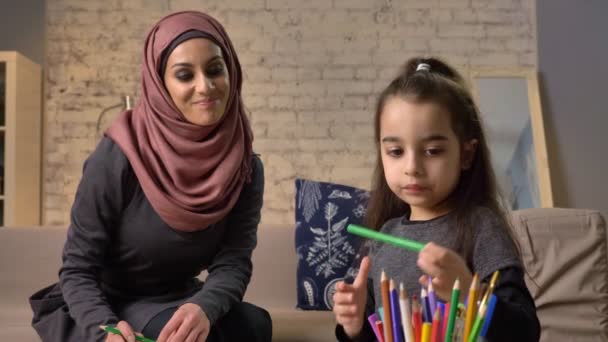 Jovem mãe bonita no hijab fazendo lições de lição de casa, pequena filha escolhe lápis de cor, desenha, coloração das crianças, conforto em casa no fundo, lápis de cor 50 fps — Vídeo de Stock