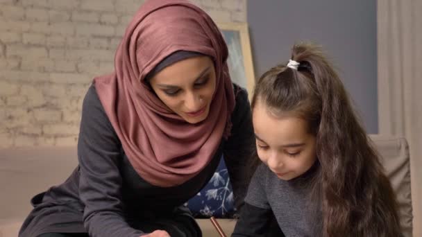 Mladá krásná matka v hidžábu dělat domácí úkoly, malá dcerka barvy se kreslí, dětské omalovánky, pastelky, domácí pohodlí v pozadí, koncepce rodinné idyly zblízka 50 fps — Stock video