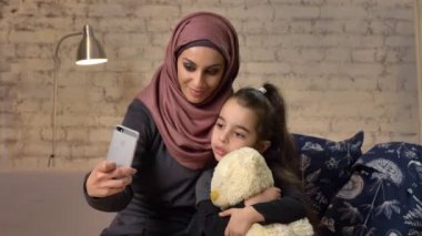Hijab kanepe, gülümseyen, kullanır smartphone, yapar selfie, sarılma, oyuncak ayı, küçük kız küçük kız ile genç güzel annem ev konforu içinde belgili tanımlık geçmiş 50 fps