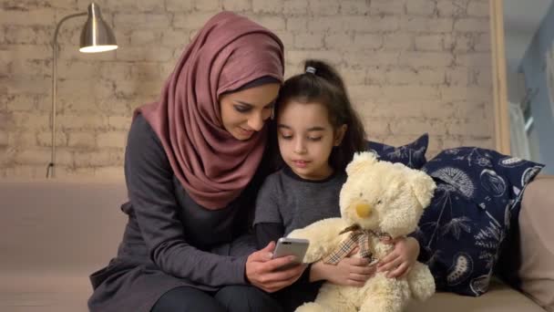 Jovem mãe bonita no hijab com a menina no sofá, sorrindo, usa smartphone, faz selfie, abraçando, menina com ursinho de pelúcia, conforto em casa no fundo 50 fps — Vídeo de Stock