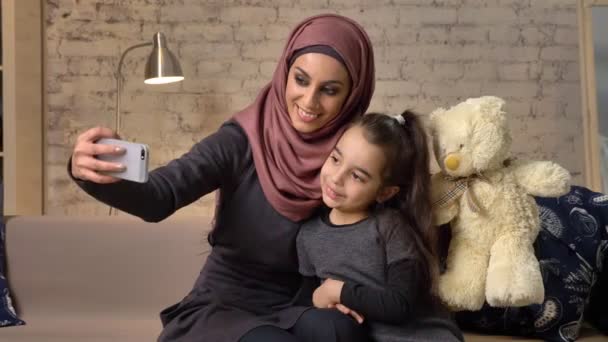 Young beautiful mother in hijab with little girl on couch, smiling, uses smartphone, makes selfie, cuddling, little girl with teddy bear, home comfort in the background 50 fps — Stock Video