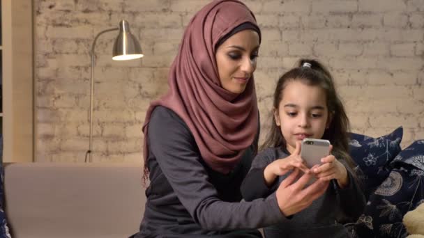 Giovane bella madre in hijab con bambina sul divano, sorridente, bambina usa uno smartphone, fa selfie, bambina con orsacchiotto, famiglia felice, concetto di comfort domestico sullo sfondo 50 — Video Stock