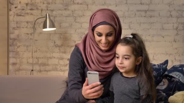 Junge schöne Mutter im Hijab mit kleinem Mädchen auf der Couch, lächelnd, nutzt Smartphone, macht Selfie, kuschelt, kleines Mädchen mit Teddybär, glückliches Familienkonzept Wohnkomfort im Hintergrund 50 fps — Stockvideo