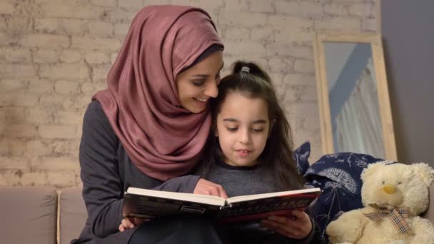 Mujer joven en hijab se sienta en el sofá con su hija y le enseña a leer, libro, concepto de familia feliz 50 fps — Vídeos de Stock