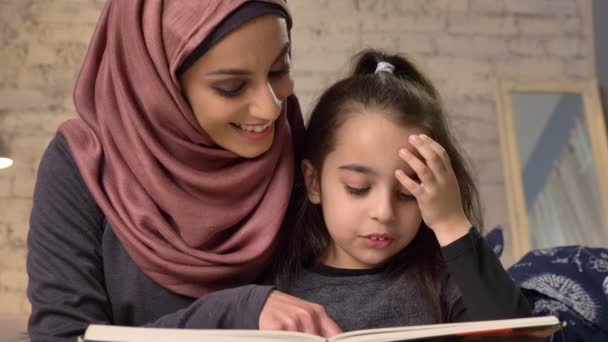 Νεαρή γυναίκα στο χιτζάμπ κάθεται στον καναπέ με την κόρη της και διδάσκει πώς να διαβάσει, το βιβλίο, ευτυχισμένη οικογένεια έννοια, κοντινό 50 fps — Αρχείο Βίντεο