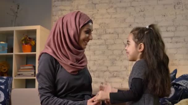 Une petite fille embrasse sa mère dans un hijab assis sur le canapé, famille, concept de confort à la maison, mère et fille. 50 ips — Video