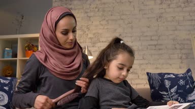 Küçük kızına oturma koltuğu, Aile, ev rahatlık kavramı, kitap okumak çocuk iken saç tarama bir hijab giyen genç Hintli bir anne kadar 50 fps kapatın