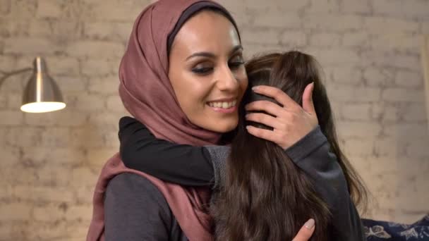 Giovane bella madre in hijab abbraccia la sua piccola figlia, guarda la fotocamera, seduta sul divano, la famiglia, sorridente, concetto di comodità a casa 50 fps — Video Stock