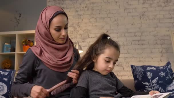 Een jonge Indiase moeder het dragen van een hijab kammen van haar naar haar dochtertje terwijl kind lezen van een boek, zittend op de Bank, familie, thuis gezelligheid concept, close-up 50 fps — Stockvideo