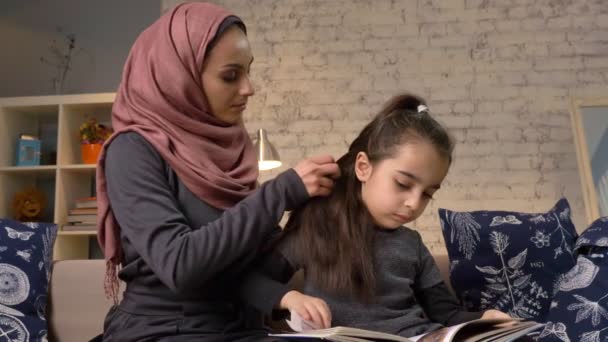 Una joven madre india que usa un hijab hace un peinado a su hija pequeña mientras que el niño lee un libro, sentado en el sofá, la familia, el concepto de comodidad en el hogar, cerca de 50 fps — Vídeos de Stock