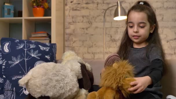 Ein kleines Mädchen sitzt auf der Couch und spielt mit einem Teddybär und einem Löwen, kämmt das Fell eines Spielzeuglöwen, Wohnkomfort im Hintergrund 50 fps — Stockvideo