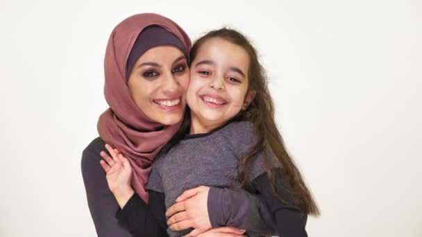 Молода красива мати в хіджабі обіймає доньку і дивиться на камеру, щаслива сім'я, концепція білого фону 50 кадрів в секунду — стокове відео
