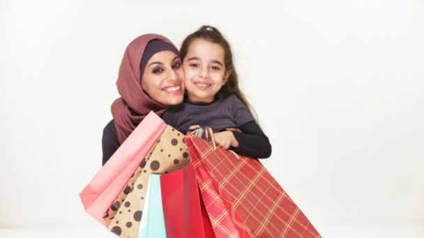 Jovem bela mãe no hijab abraça sua filhinha, segurando sacos de compras, sorrindo, família feliz no fundo branco, conceito 50 fps — Vídeo de Stock