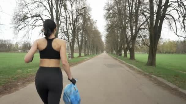 Chica atlética joven en un traje deportivo negro corre a través del verde parque de verano con una bolsa de basura en la mano, concepto de trote, vista trasera 50 fps — Vídeo de stock