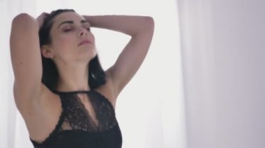 Genç seksi beyaz esmer siyah iç çamaşırı kollarını yukarı yükseltir, saçlarını dokunur, belgili tanımlık pencere, gün ışığından yararlanma yakınındaki gözlerini kapamaz 50 fps