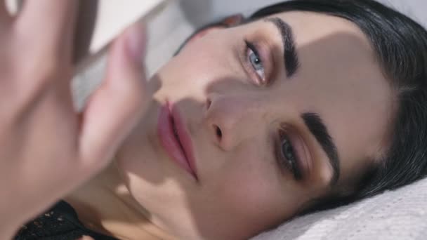 Close-up retrato de uma jovem deitada em um travesseiro e usando um smartphone, morde o lábio, luz do dia, feixe de 50 fps — Vídeo de Stock