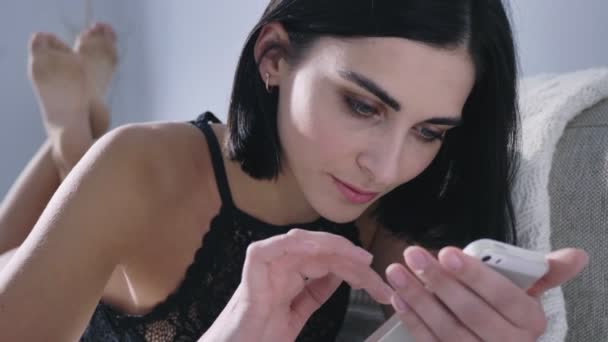 Jeune brune caucasienne sexy utilisant un smartphone, défilant, allongée sur le canapé, jambes sexy en arrière-plan, lumière du jour, gros plan 50 images par seconde — Video