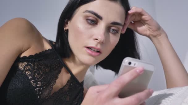 Mladá sexy kavkazské bruneta v černém prádle pomocí smartphone, ležící na pohovce, usměvavý, denní světlo, pokušení, zblízka 50 fps — Stock video