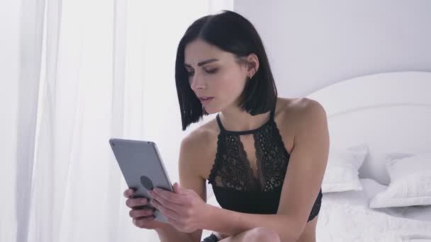 Jeune fille brune caucasienne sexy en lingerie noire à l'aide d'une tablette, regard mécontent, fond de chambre blanche 50 images par seconde — Video