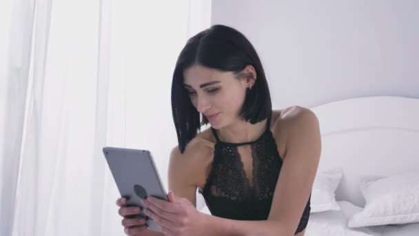 Jonge sexy Kaukasische brunette meisje in zwarte lingerie met behulp van een tablet, lachen, videochat concept, wit slaapkamer achtergrond 50 fps — Stockvideo