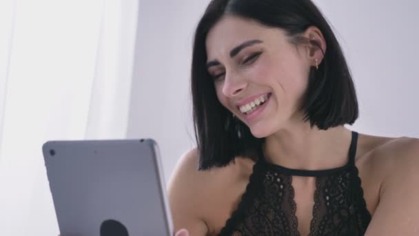 Giovane ragazza caucasica sexy bruna in lingerie nera utilizzando un tablet, ridere, sorridente, concetto di video chat, sfondo della camera da letto bianca, primo piano 50 fps — Video Stock