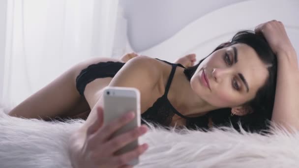 Ung sexig kaukasiska brunett tjej i svarta underkläder med en smartphone, gör selfie, ligger på mage, fluffig filt, vita sovrum bakgrunden 50 fps — Stockvideo