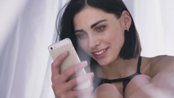 Retrato de una joven morena caucásica sexy en lencería usando lectura de teléfonos inteligentes, sonriente, fondo blanco 50 fps — Vídeos de Stock