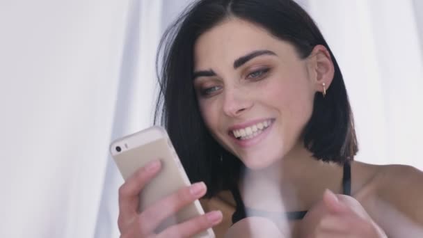 Portret van jonge sexy Kaukasische brunette meisje in lingerie met behulp van de smartphone, lezen, glimlachen, lachen, wachtruimten hoofd met hand, witte achtergrond 50 fps — Stockvideo