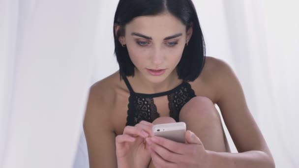 Portrait of young sexy caucasian brunette girl in lingerie using smartphone reading, swiping, holds head with hand, white background 50 fps — Stock Video