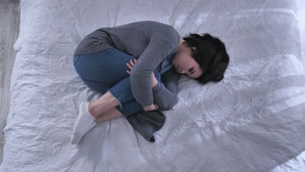 Молодая грустная азиатка лежит в позе эмбриона на кровати, держа колени руками, несчастная, белая спальня 50 кадров в секунду — стоковое видео