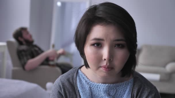 Porträtt av en ledsen deprimerad asiatisk flicka, berusad man i bakgrunden sitter i en fåtölj, ser in i kameran. 50 fps — Stockvideo