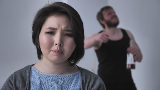 Portrait of a sad depressed Asian girl, drunk husband in the background swears, quarrel, conflict, looks at the camera. 50 fps — Stock Video