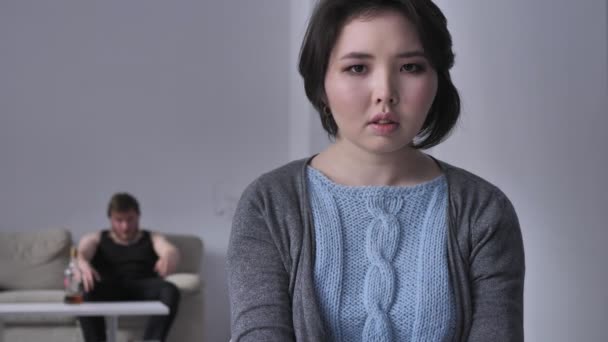 Portrét smutné depresivní asijské dívky, opilý manžel v pozadí sedí na pohovce, pití alkoholu, násilí, při pohledu na fotoaparát. 50 fps — Stock video