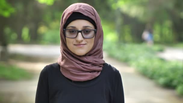 Portrait of a young smiling girl in glasses wearing hijab, outdoor, in a park in the background. 50 fps — Stock Video