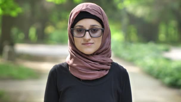 Портрет молодой смеющейся девушки в очках в хиджабе, на открытом воздухе, в парке на заднем плане. 50 кадров в секунду — стоковое видео