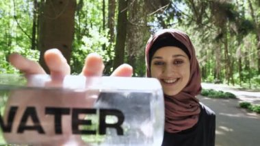 Bir hijab bir sevimli genç kız portresi su gülümseyerek, kamera, arka plan, parkta bakarak onun elinde bir şişe ile odak 50 fps çekin