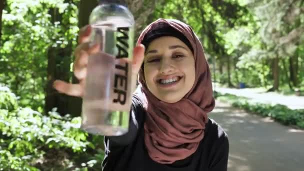 Portrait of a cute young girl in a hijab with a bottle of water in her hands, smiling, looking at the camera, park in the background, focus pull 50 fps — Stock Video