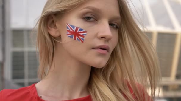 Portrét mladé blond dívka fotbalový fanoušek z Anglie do kamery, vážnou tvář, stadion v pozadí 50 fps — Stock video