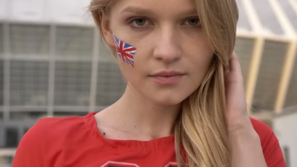 Retrato de una joven chica rubia fanática del fútbol de Inglaterra mirando a la cámara, cara seria, estadio en el fondo 50 fps — Vídeos de Stock